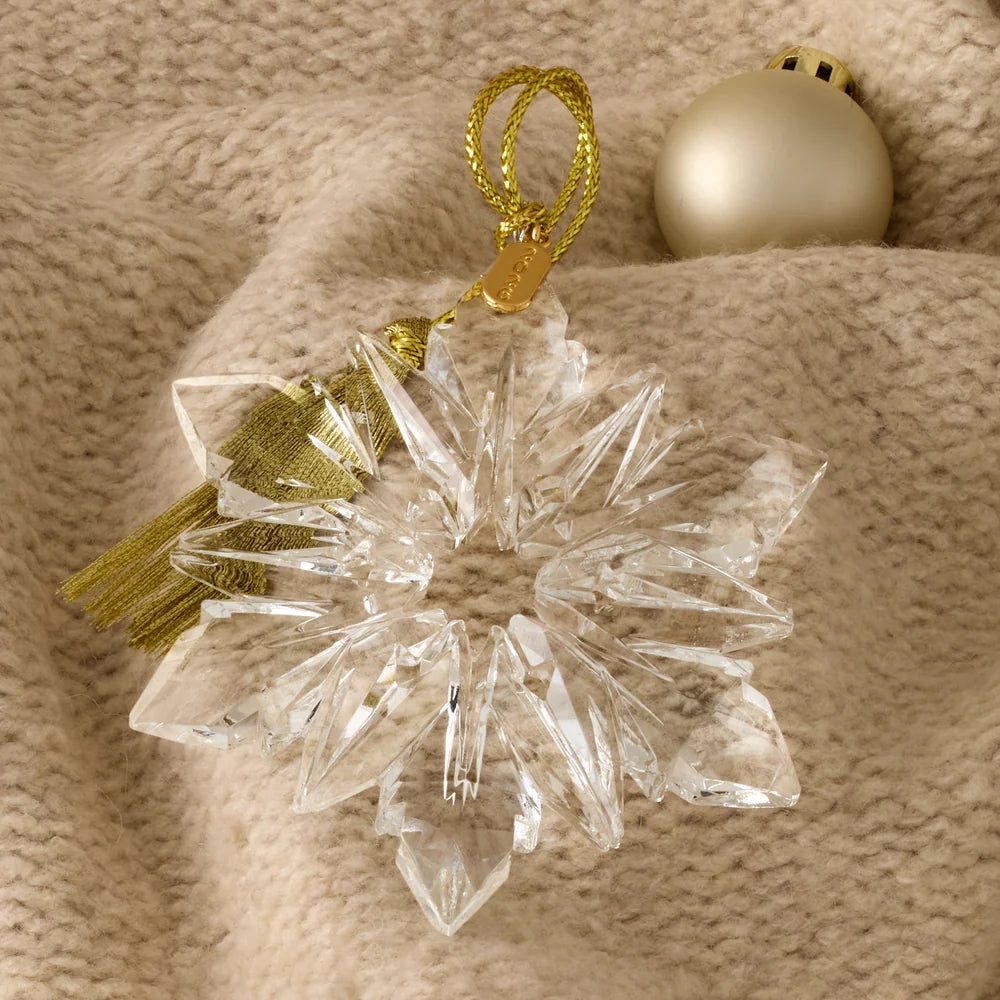 A clear faceted crystal snowflake ornament with a gold hanging loop and tassel, displayed on a beige knit blanket next to a gold Christmas bauble.