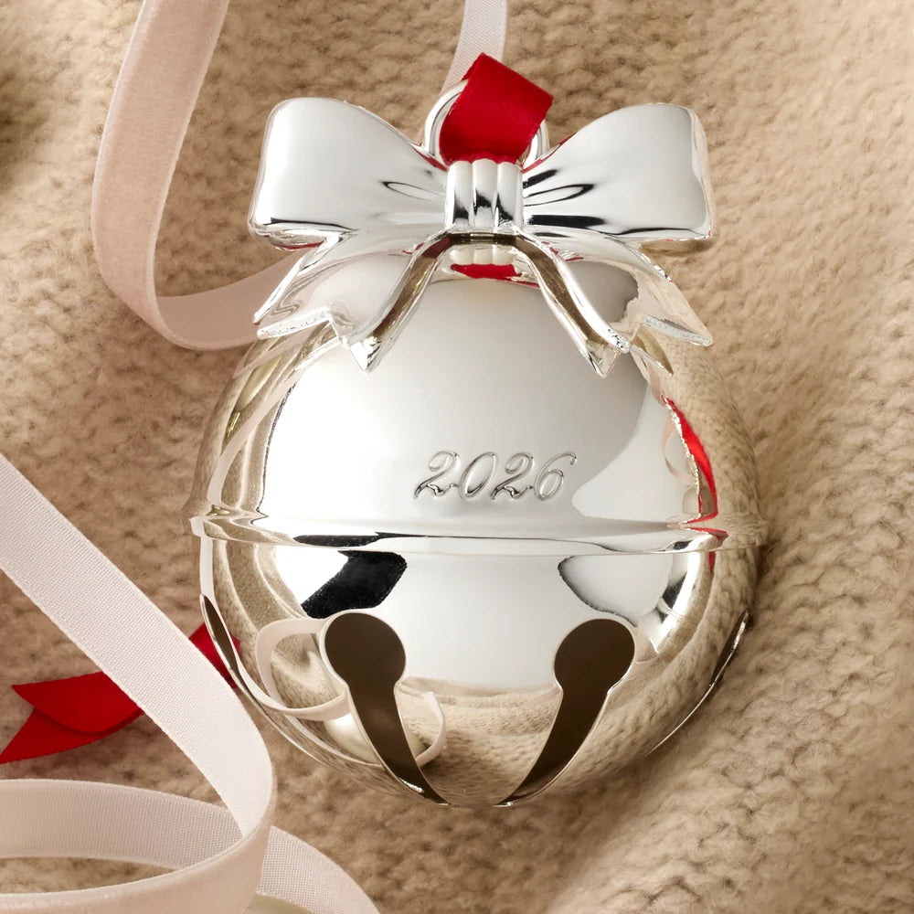 A shiny silver Christmas bell ornament engraved with the year 2026, decorated with a silver bow and red ribbon, resting on a beige textured fabric background.