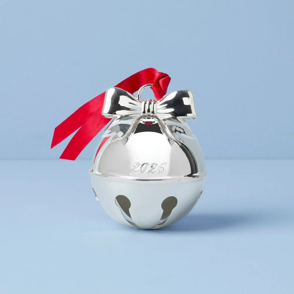 A shiny silver Christmas bell ornament with a red satin ribbon bow and a '2026' engraving, set against a light blue background.