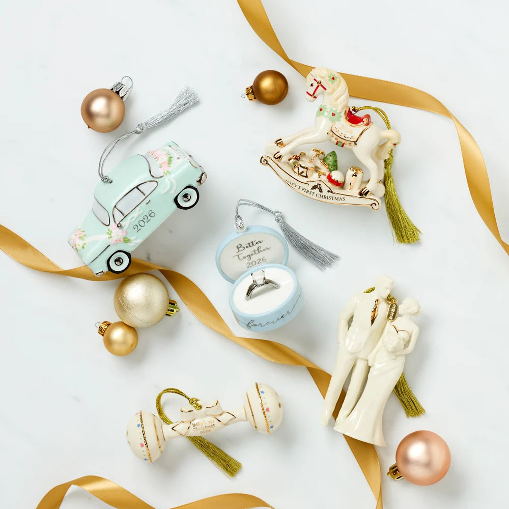 A styled flat-lay of Christmas ornaments on a white background with gold ribbons and gold and rose gold baubles. The ornaments include a mint green car, a rocking horse, a ring box with an engagement ring, a couple kissing, a baby carriage, and a small Santa ornament.