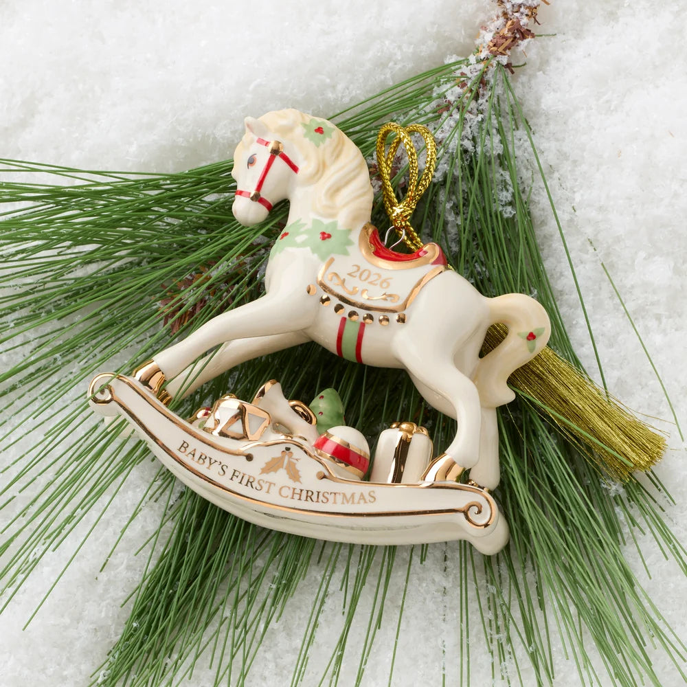 A ceramic Christmas ornament shaped like a rocking horse carrying a sleigh with gifts, placed on green pine branches over a white snowy background. The ornament is cream-colored with gold trim, red accents, and the text '2026 Baby's First Christmas' and 'Baby's First Christmas'.