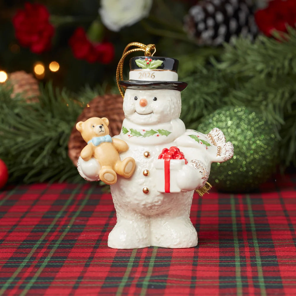 A ceramic Christmas snowman ornament stands on a red plaid surface with pine branches, ornaments, and warm lights blurred in the background. The snowman is white with gold buttons, a black top hat with green holly accents, and holds a small beige teddy bear and a white gift box with red ribbon.