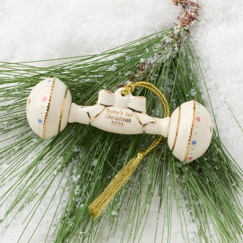 Multicolored baby rattle ornament with "Baby's 1st Christmas 2026" written in gold on it, shown on pine needles and a snowy background