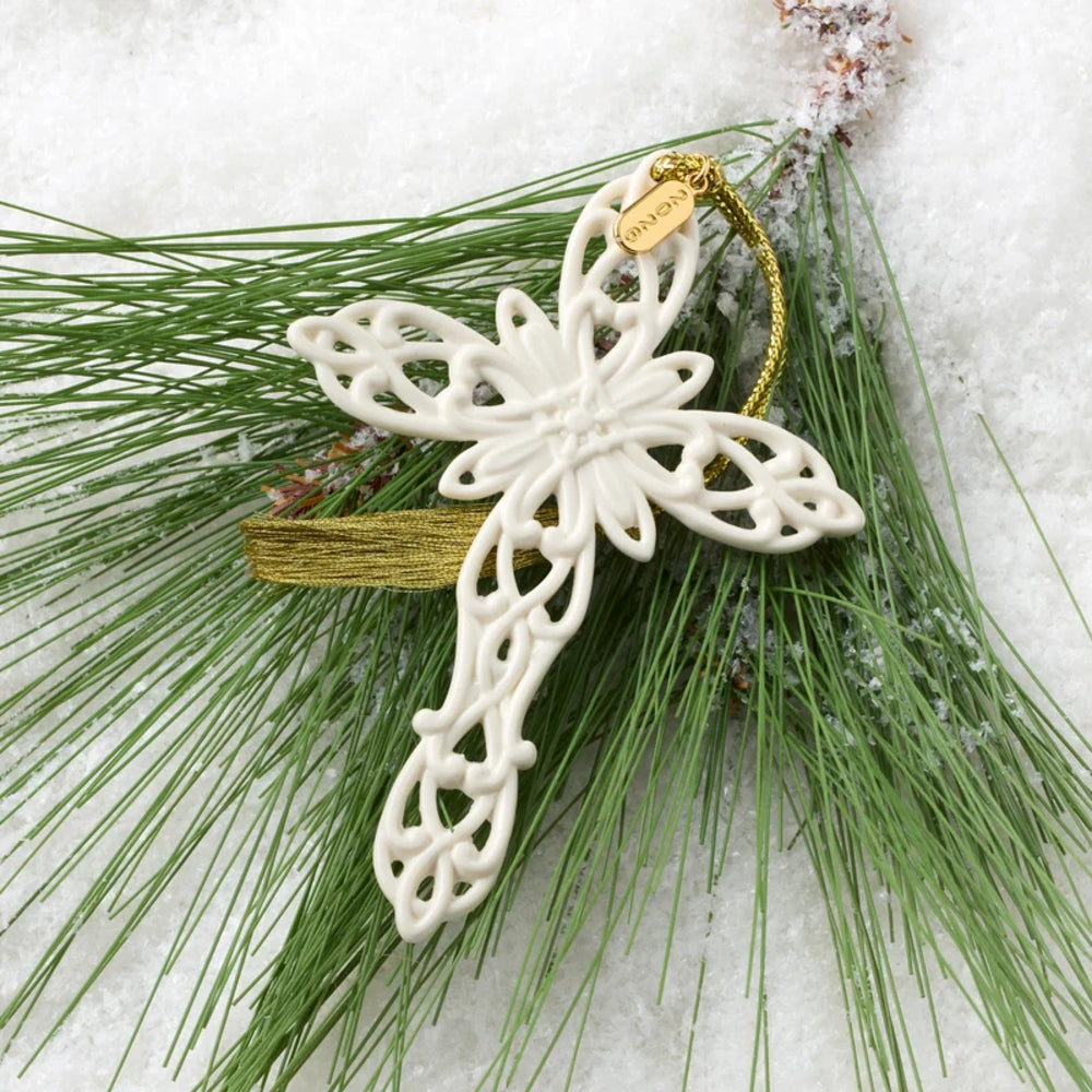 An off-white ceramic Christmas ornament shaped like a stylized cross with intricate cutout details, hanging from a gold string. It is styled on green pine needles with light snow dusting, against a white snowy background.
