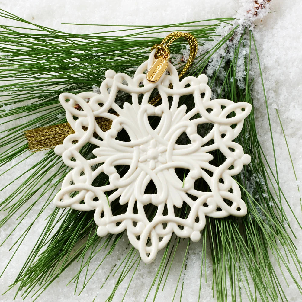 An off-white ceramic snowflake Christmas ornament with a gold hanging loop, displayed on green pine needles with a dusting of snow.