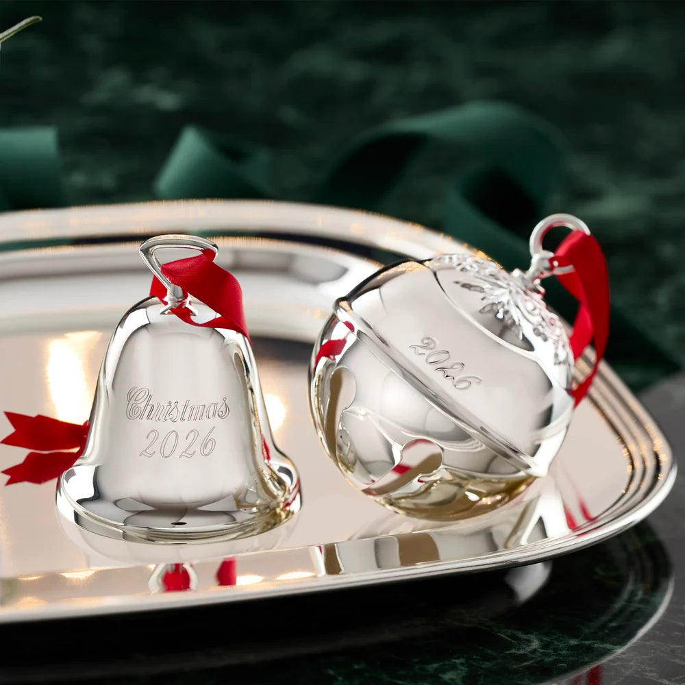 Two silver bells with red ribbons on a reflective surface with a blurred green background