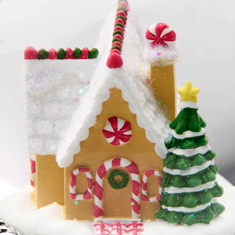 A festive gingerbread-style house decoration or cake topper with a white glittery snow-covered roof, red and green candy accents, a candy cane window, a green wreath on the door, and a small green Christmas tree beside it on a white background.