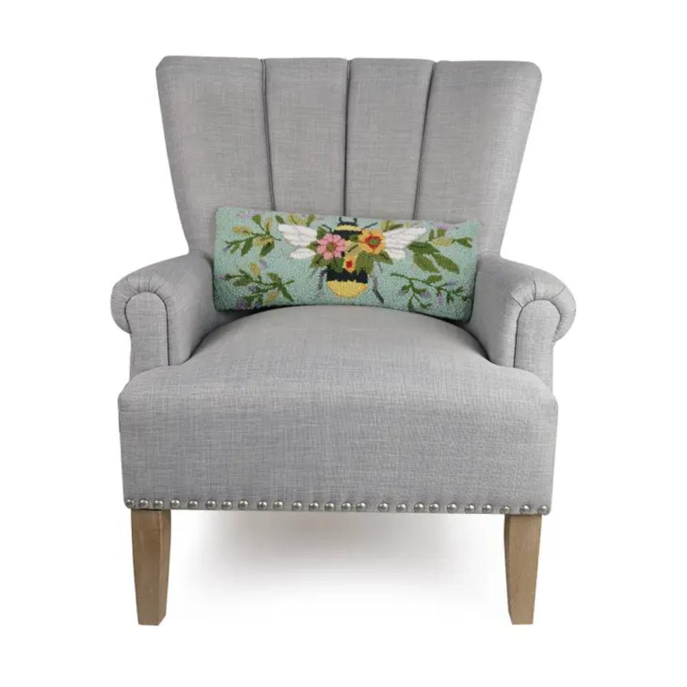 A rectangular accent pillow on a chair with a floral pattern and a bee design in the center, predominantly in shades of green, blue, and yellow.