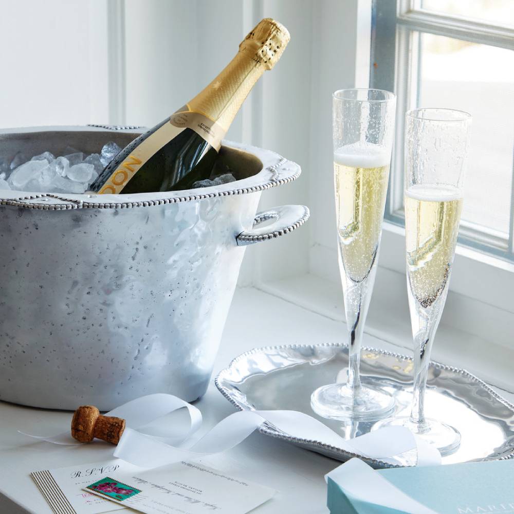 Bottle of champagne in an ice bucket with two glasses on a tray by a window.