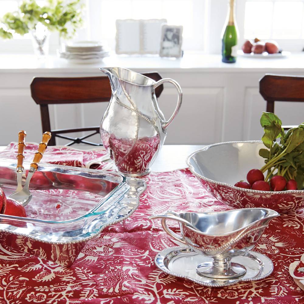 Dining table set with silverware and a pitcher on a red patterned tablecloth.