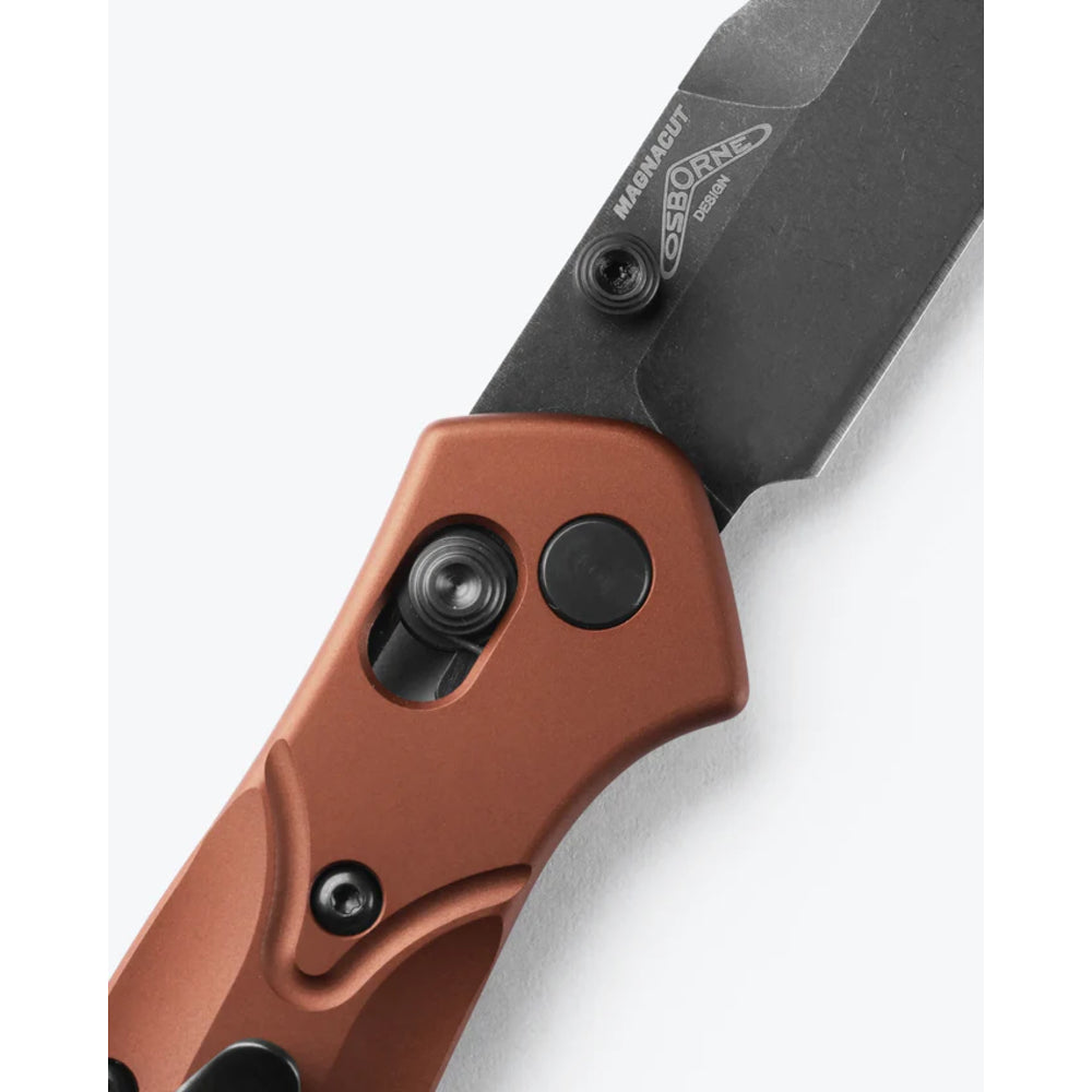Close-up of a folding knife with a matte black blade and a bronze-finished handle, showing the Magellan brand logo on the blade.