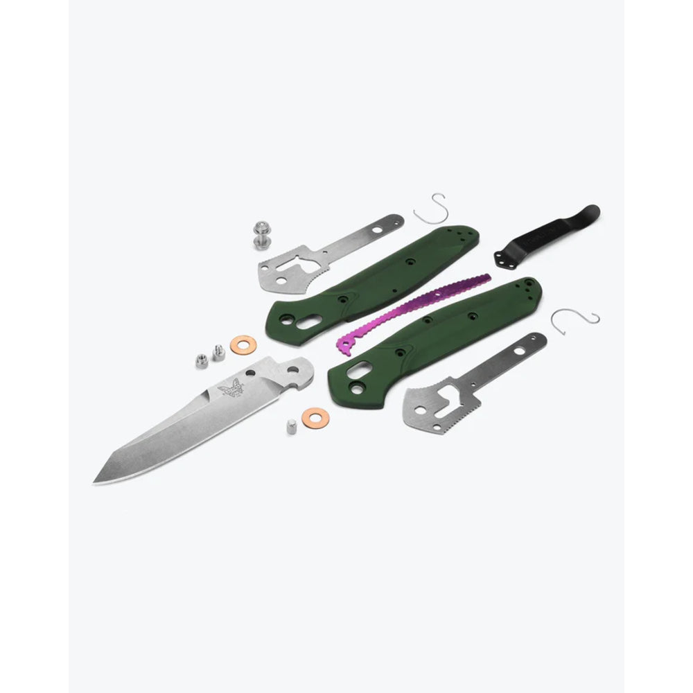 A knife blank kit with a partially fabricated silver blade, green handle scales, metal tangs, a black handle wrap, a purple lanyard, and small hardware pieces arranged on a light gray background.