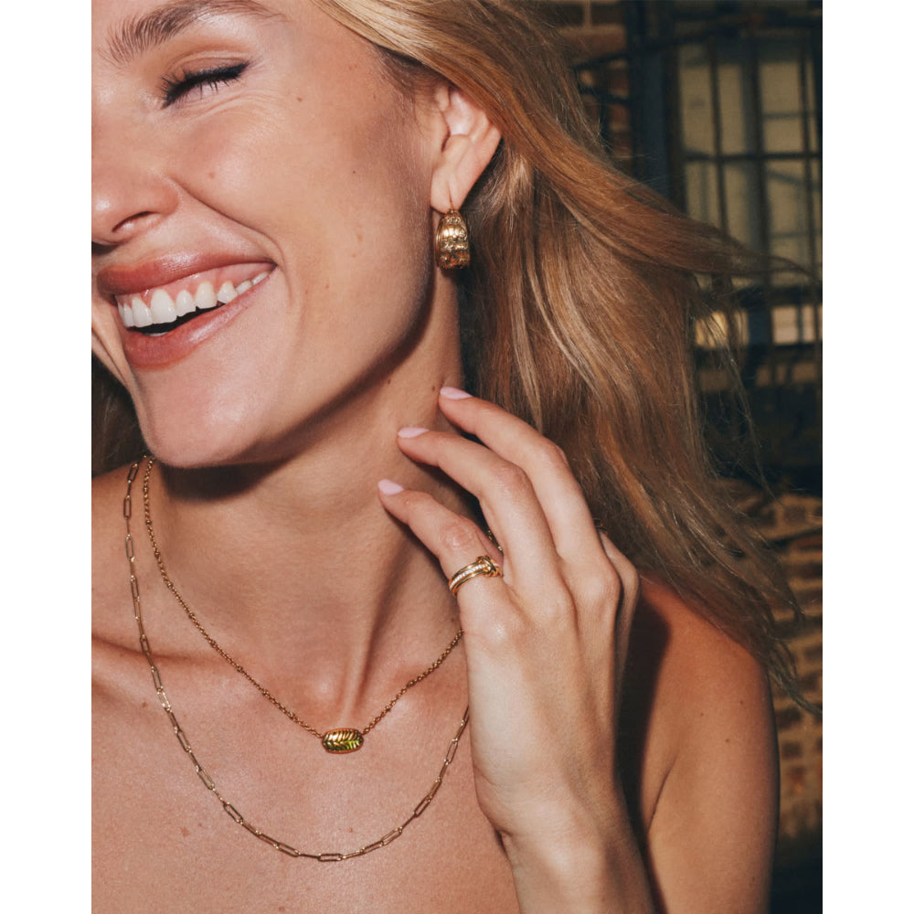 A close-up lifestyle shot of a smiling woman wearing gold jewelry, including a delicate chain necklace with a small gold bead, a matching ring, and a gold hoop earring, against a warm-toned indoor background.