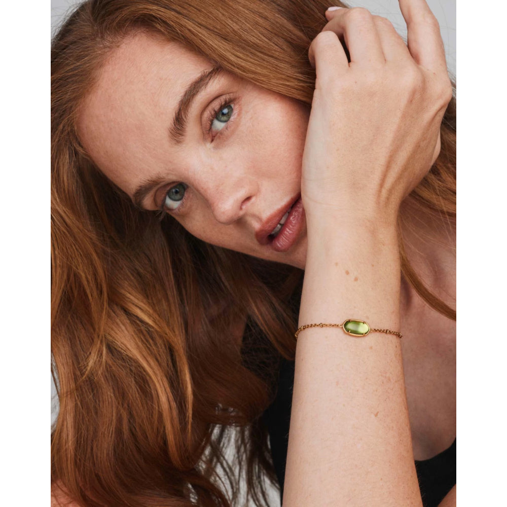 A close-up lifestyle shot of a woman with long wavy auburn hair wearing a delicate gold chain bracelet with a small, polished olive-green gemstone on her wrist.