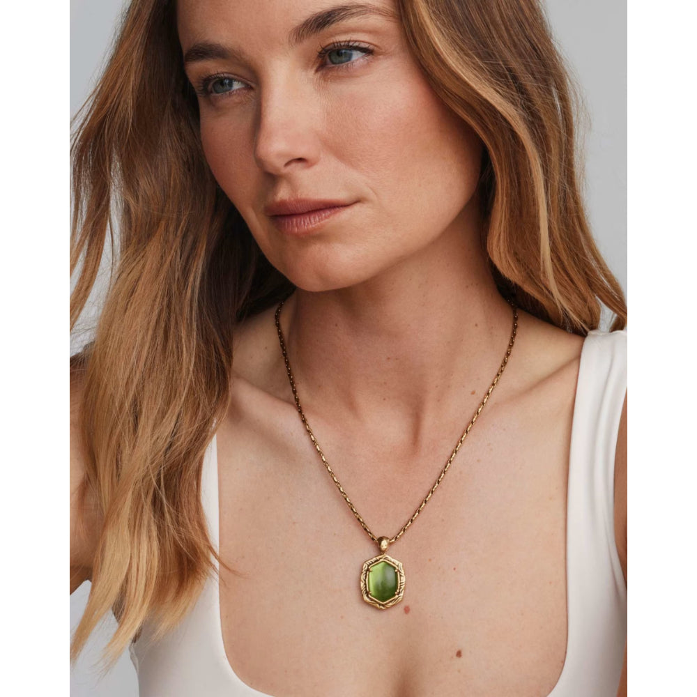 A woman wearing a delicate gold chain necklace with an oval green pendant, shown in a close-up portrait against a light gray background.