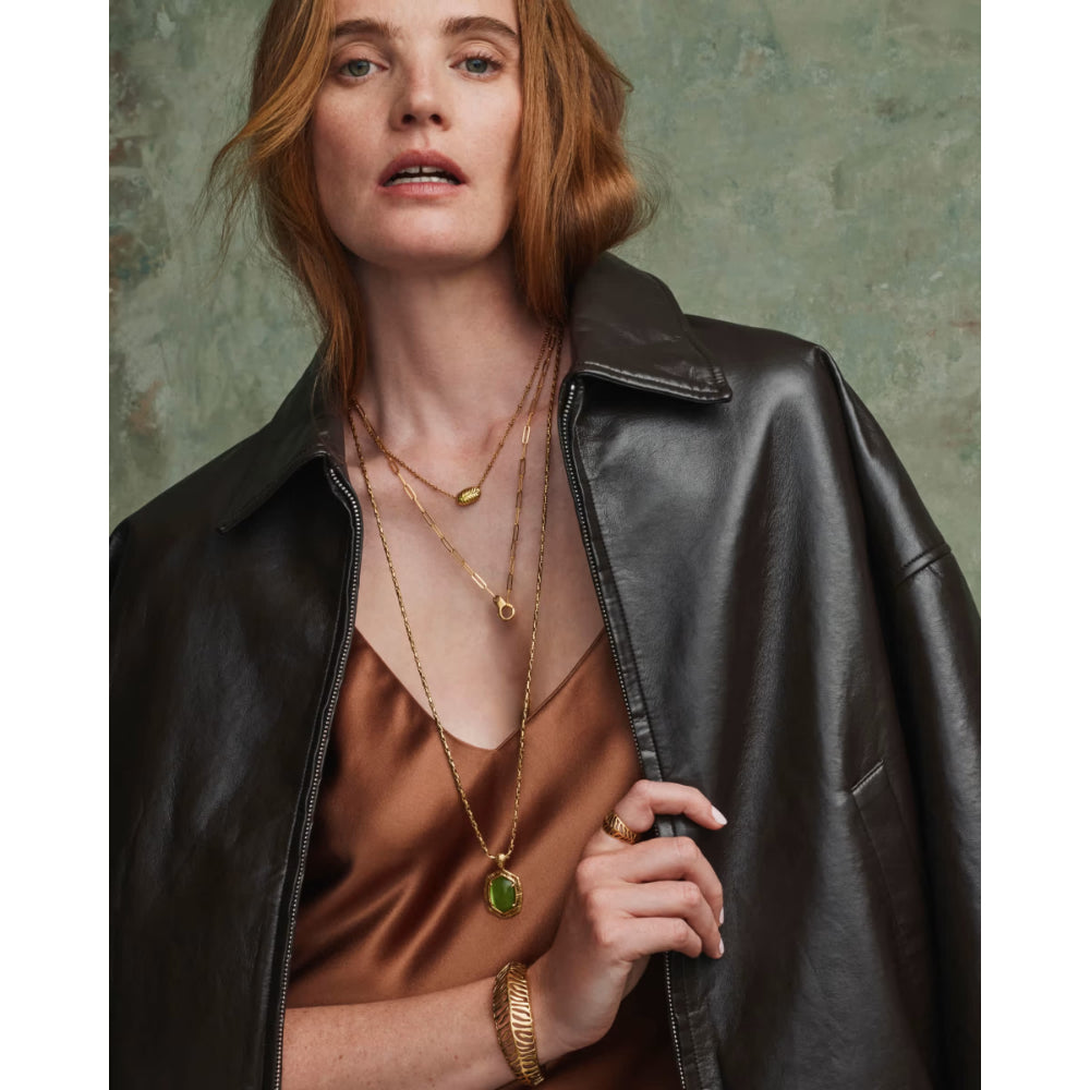A woman wearing a black leather jacket over a bronze satin top, accessorized with layered gold necklaces, a green pendant, and a gold cuff bracelet, posed against a textured green background.