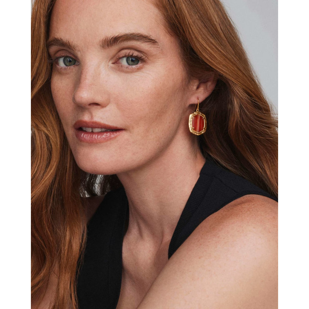 A close-up portrait of a woman with long wavy light brown hair wearing a gold-framed rectangular drop earring with a red-orange stone, against a light gray background.