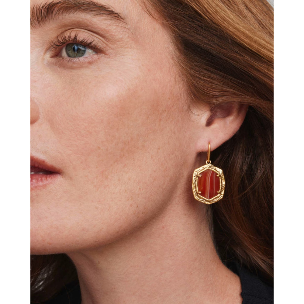 A close-up of a woman's ear wearing a gold drop earring with a faceted red-orange stone.