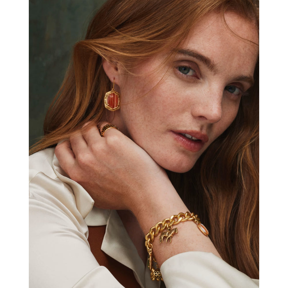 A close-up lifestyle shot of a woman with red hair wearing a gold chain bracelet with a small gold star charm, a gold ring, and a gold drop earring with a red-orange geometric inset.
