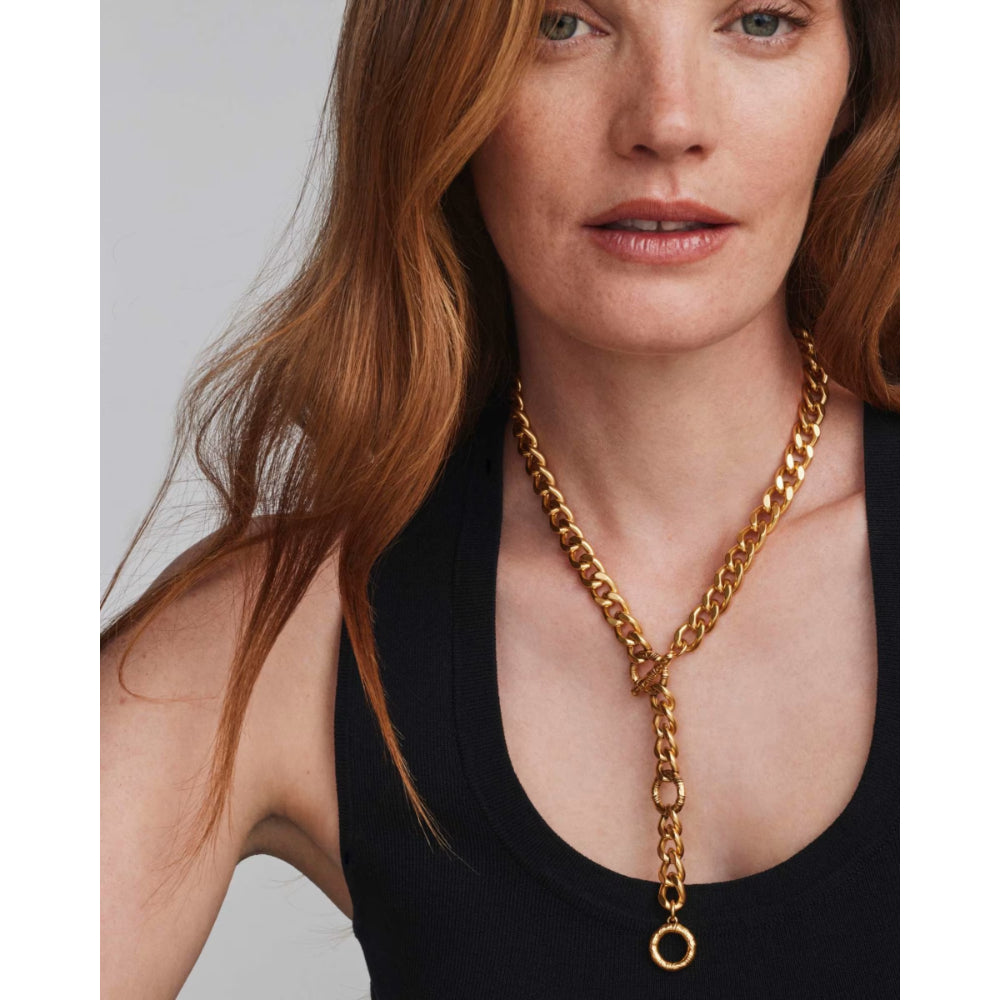 A close-up of a woman with red hair wearing a chunky gold chain necklace with a Y-shaped design and a hanging ring pendant, paired with a black tank top against a light gray background.