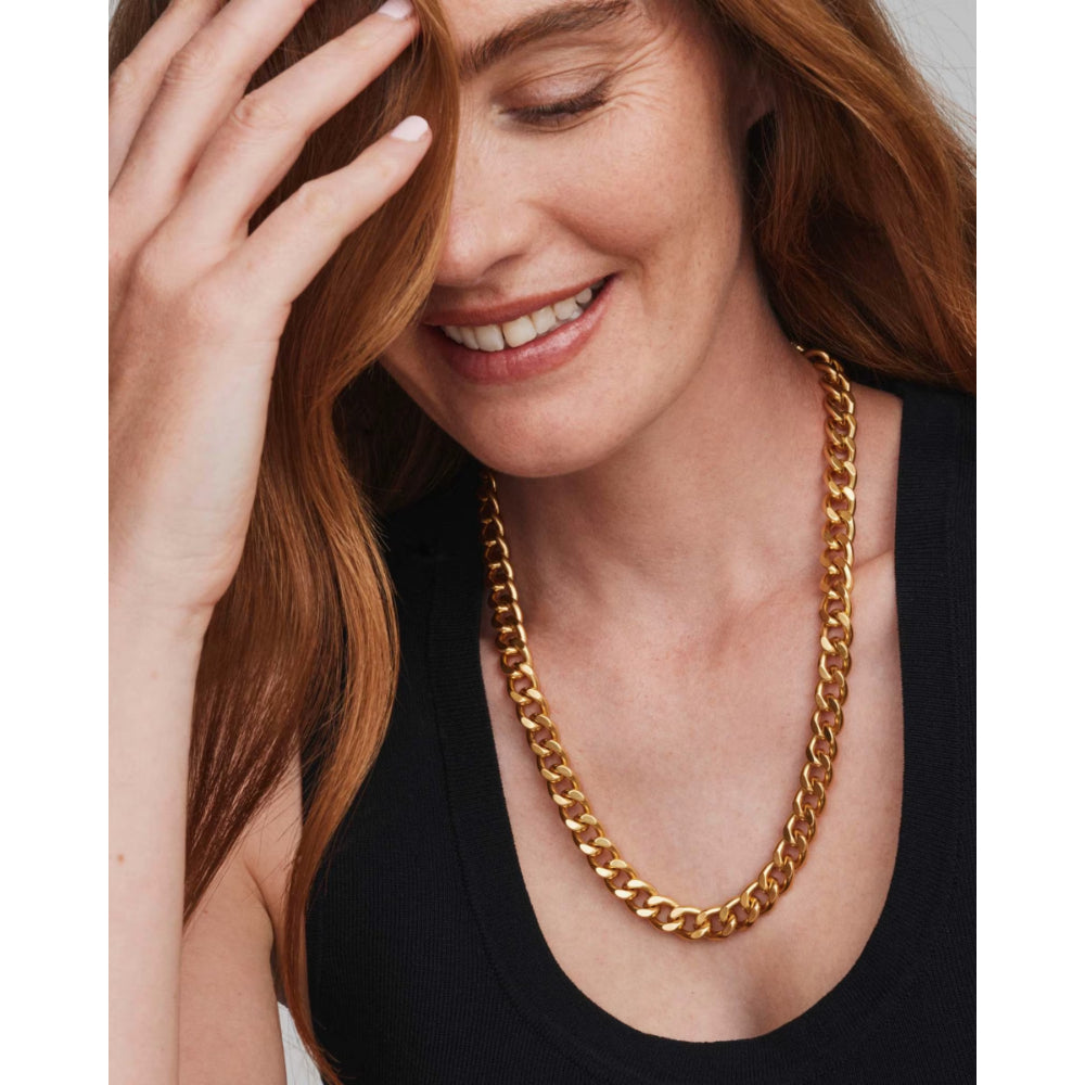 A close-up of a woman with long red hair wearing a chunky gold chain necklace and a black tank top, with one hand touching her face.