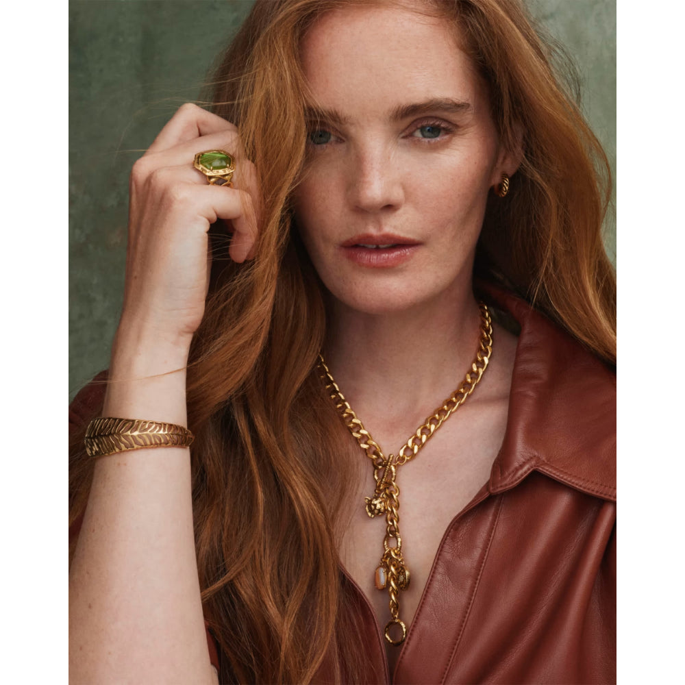 A close-up portrait of a woman with long red hair wearing a chunky gold chain necklace with a dangling charm, a gold ring, and a gold bracelet, paired with a brown leather jacket against a textured green background.