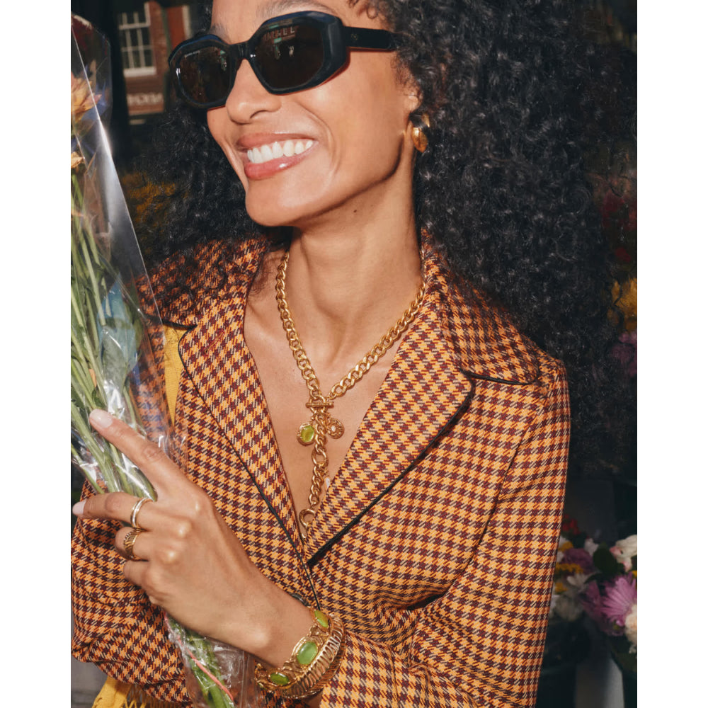 A woman wearing a brown and black houndstooth blazer, gold chain necklaces, sunglasses, and holding a bouquet of flowers.