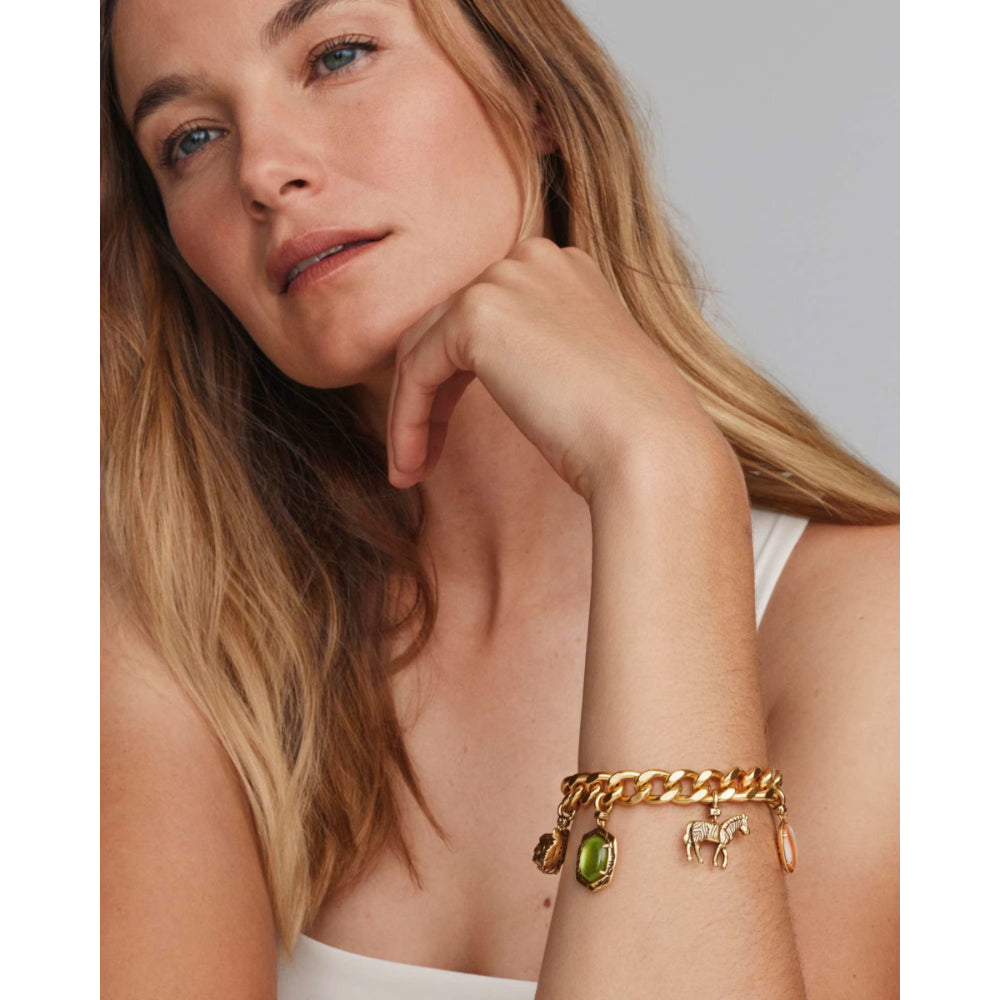 A close-up of a woman wearing a gold chain charm bracelet with a green oval stone and a horse charm, set against a neutral gray background.