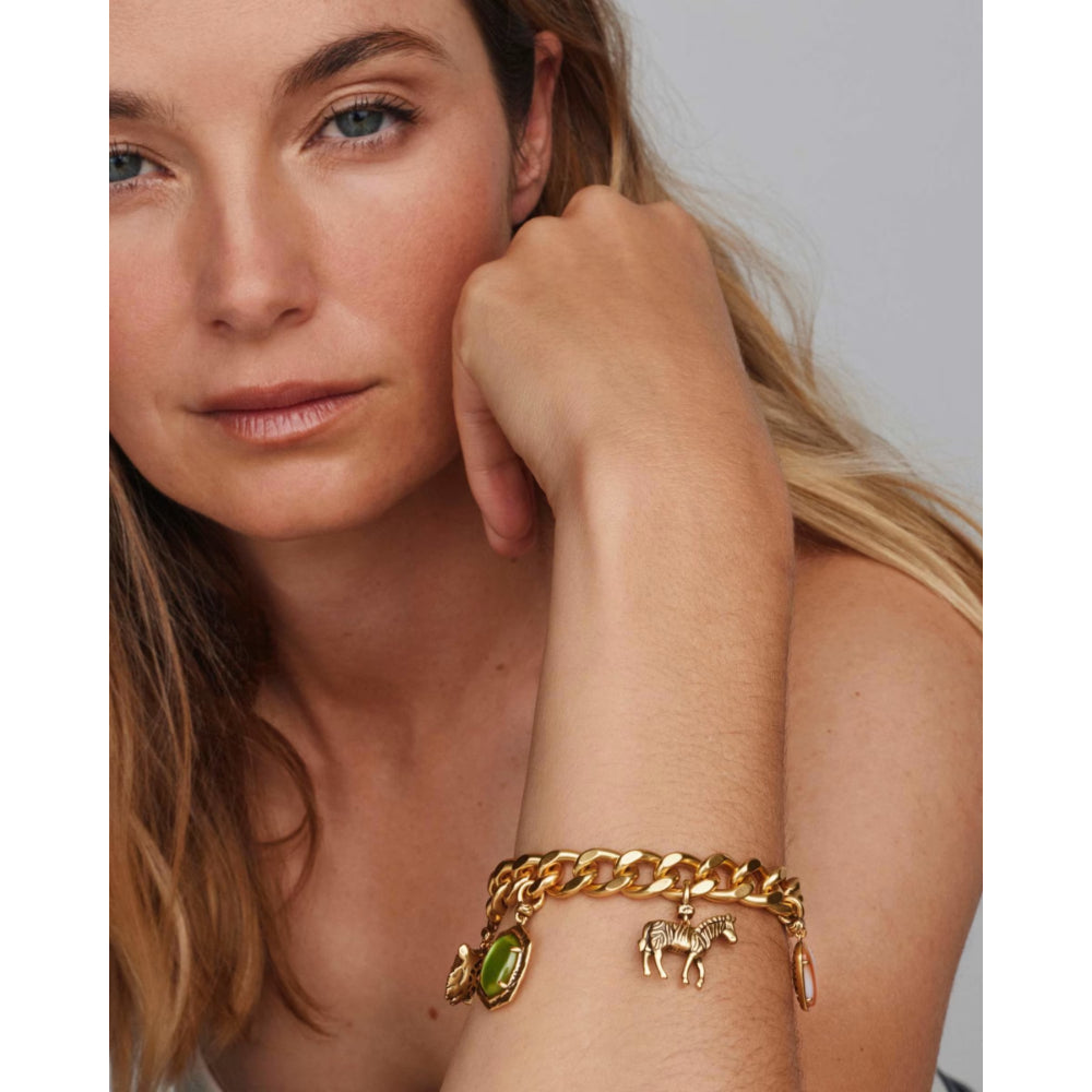 A close-up of a woman wearing a gold chain bracelet with horse charms and a green gemstone charm on her wrist, set against a neutral gray background.