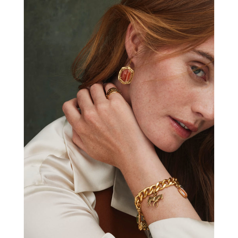 A close-up lifestyle shot of a woman wearing gold jewelry, featuring a chunky gold chain bracelet with a small gold charm, a gold ring, and a gold geometric earring with a red-orange stone.