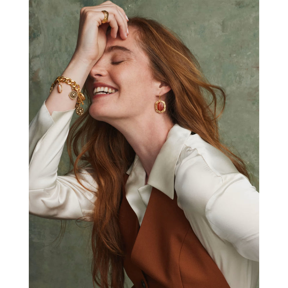 A woman with long red hair wearing gold jewelry, including a gold chain bracelet with charms on her wrist and matching gold drop earrings, posing against a textured gray-green background.