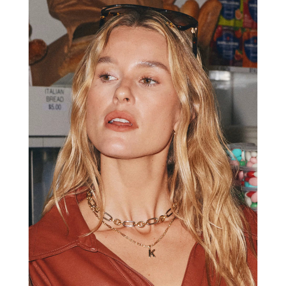 A close-up lifestyle shot of a blonde woman wearing layered gold necklaces, one with a small 'K' initial pendant, in a cafe setting.