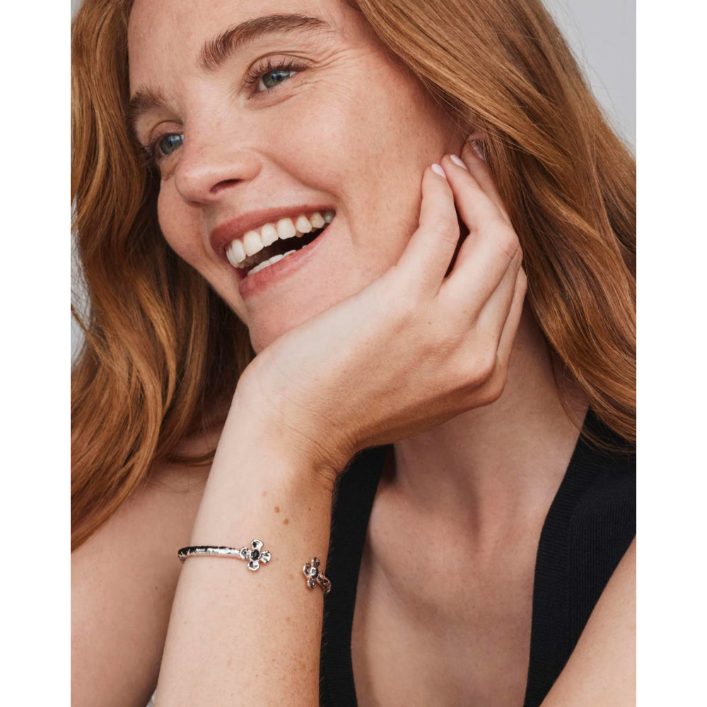 A close-up lifestyle shot of a smiling woman with red hair wearing a delicate silver bangle bracelet on her wrist, featuring small star and flower charms.