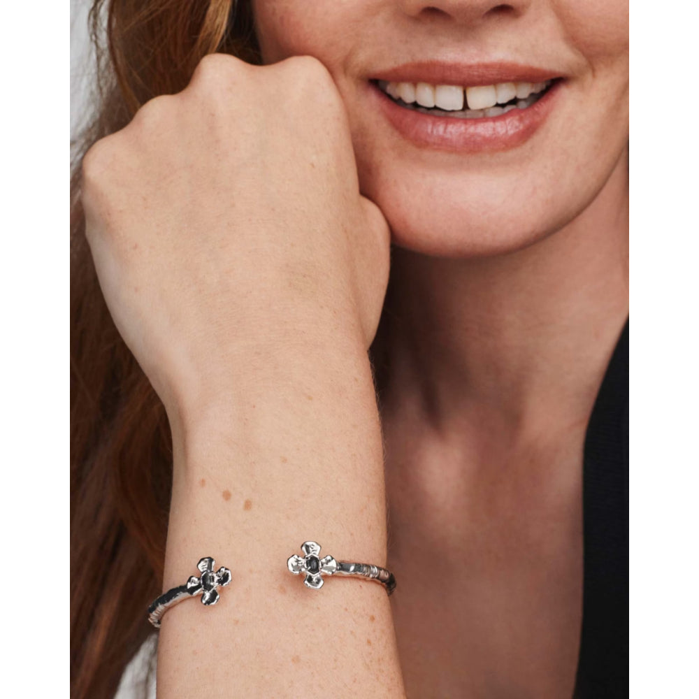 A close-up of a woman's wrist wearing two silver-toned bangle bracelets, each featuring a small floral or clover-like motif with clear stones. The photo is cropped to show the jewelry on the model's wrist, with part of her face and hand visible.