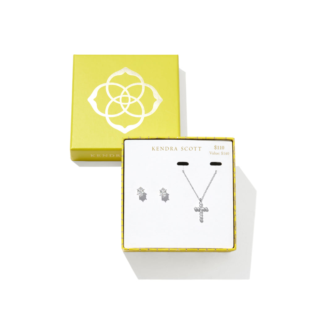 Kendra Scott silver jewelry set featuring a cross pendant necklace and matching stud earrings, displayed in a white presentation box with gold trim, next to its lime green Kendra Scott packaging box on a white background.