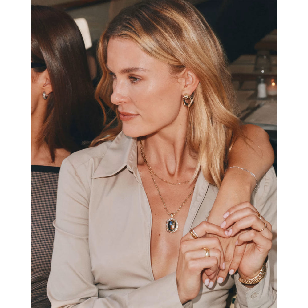 A blonde woman in a beige button-up shirt is shown in a social setting, wearing layered gold necklaces, rings, and bracelets.