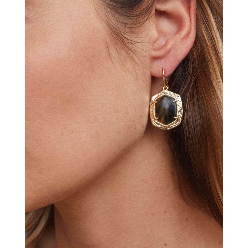 A close-up of a model's ear wearing a textured gold drop earring with a dark, polished tiger's eye stone.