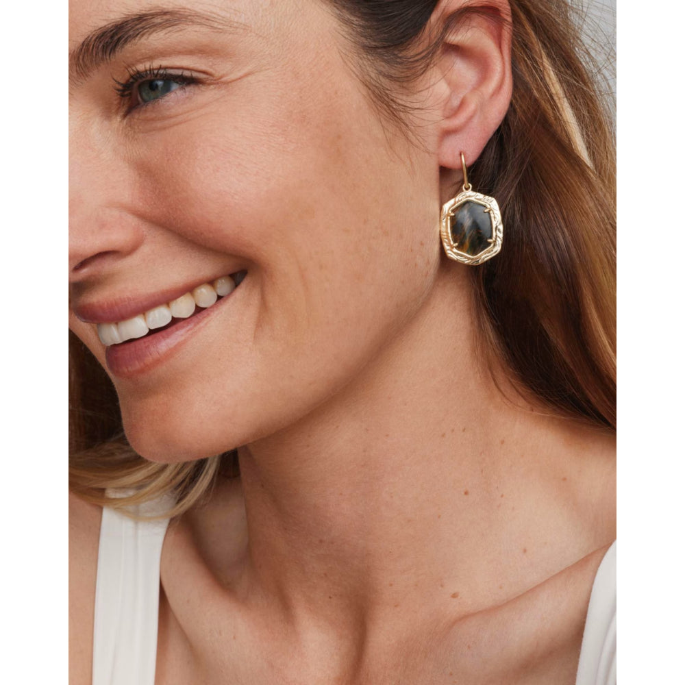 A close-up of a woman wearing a gold geometric drop earring with a dark gray stone, shown against her ear and hair.