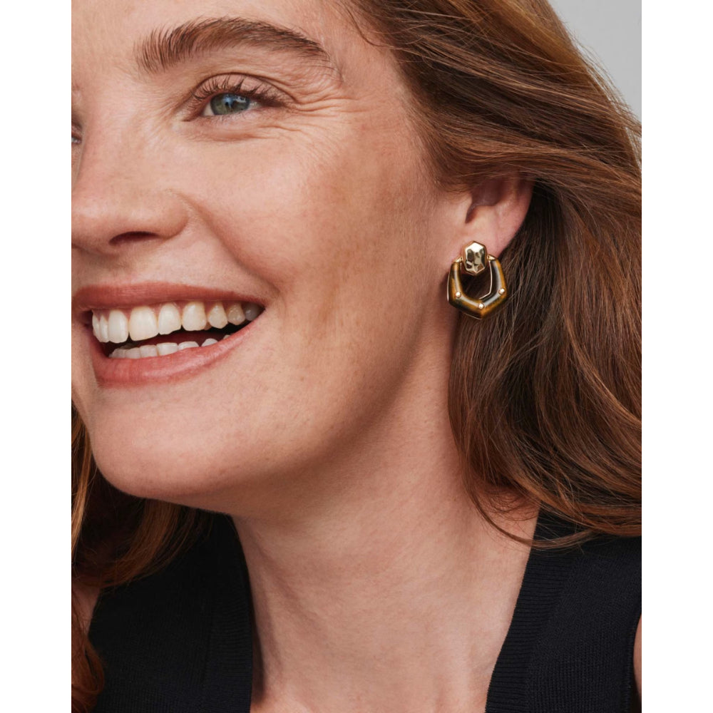 A close-up of a smiling woman wearing a gold geometric hoop earring with a small clear stone accent.