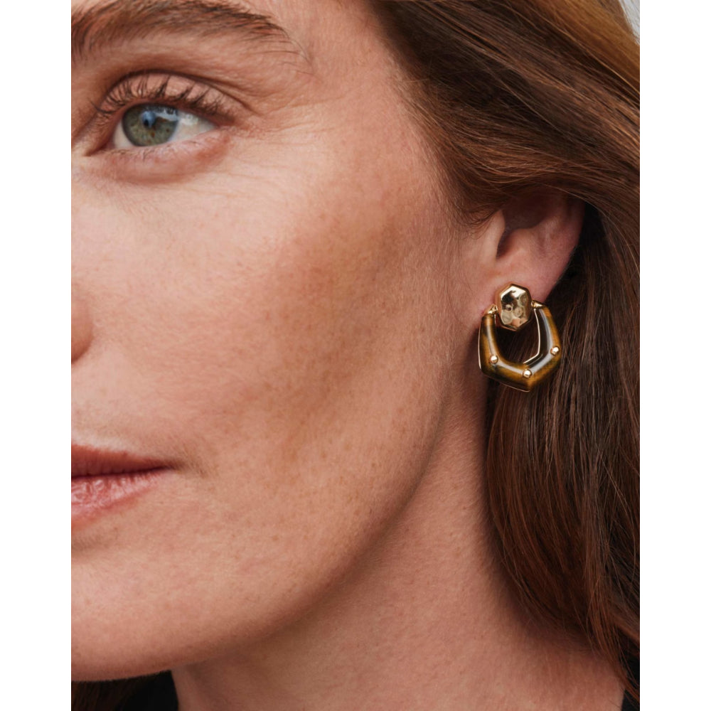 A close-up of a woman's ear wearing a gold-toned geometric hoop earring with a small skull detail at the top.