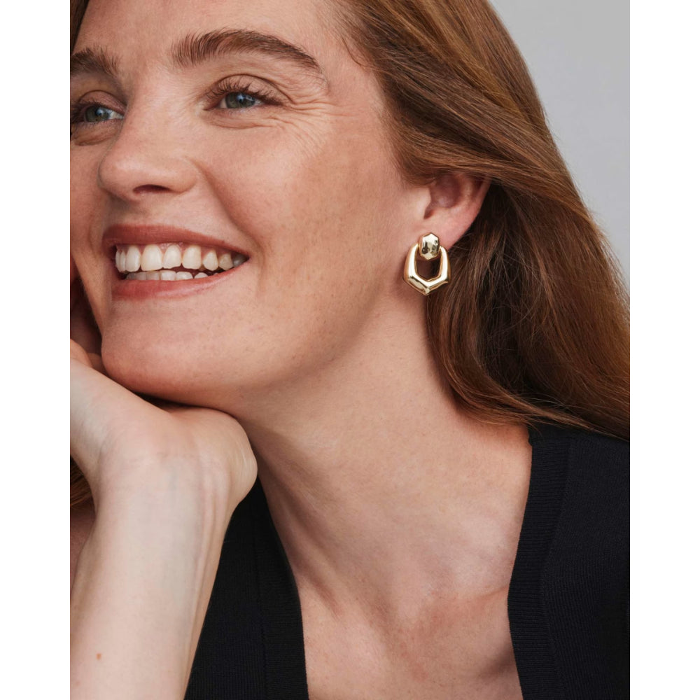 A close-up of a smiling woman with brown hair wearing a gold geometric hoop earring, resting her chin on her hand against a neutral gray background.