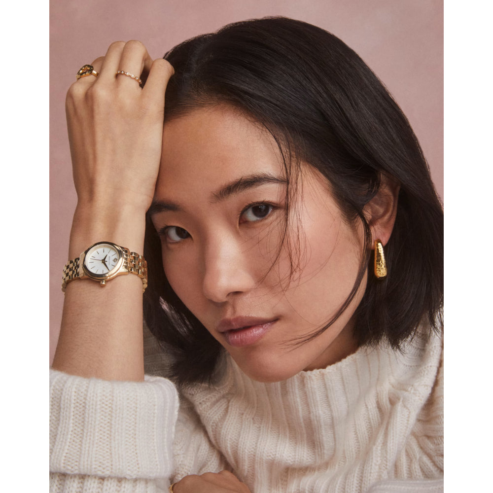 A close-up lifestyle shot of a woman with short dark hair, wearing a gold-tone wristwatch with a white dial and a gold chain bracelet, along with gold hoop earrings and rings, set against a solid dusty pink background.