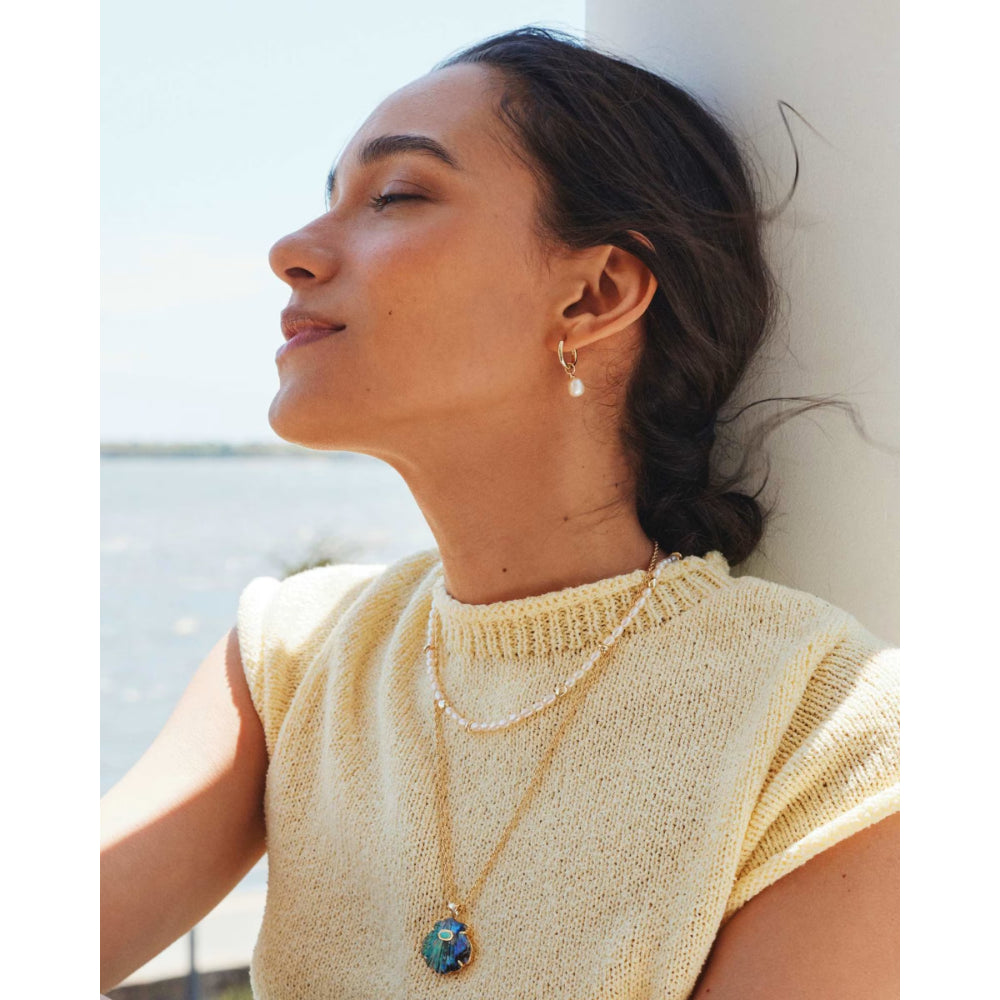 Woman wearing a yellow knit top and gold necklace with a blue pendant, leaning against a white wall.