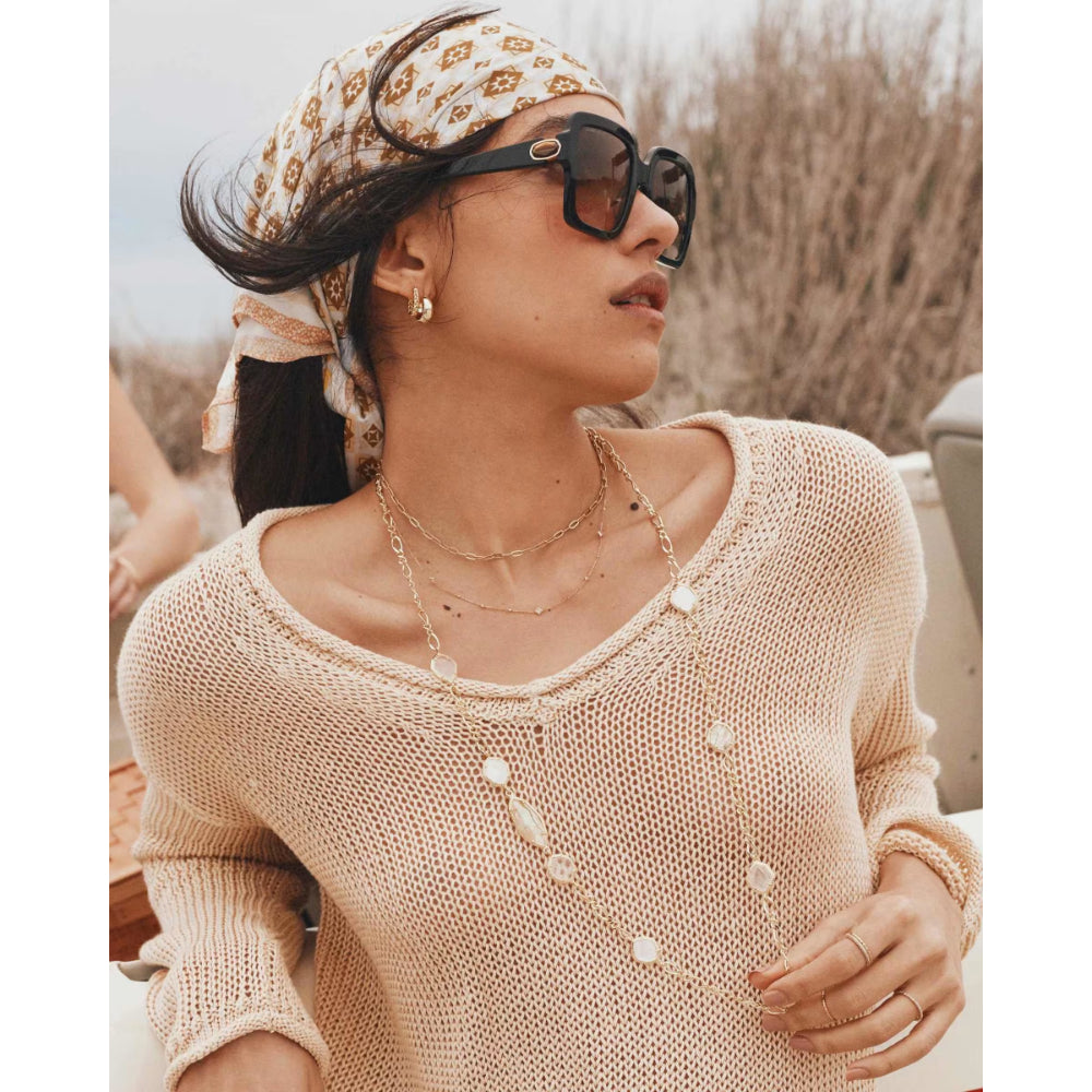 Woman wearing a beige knitted sweater, sunglasses, and a patterned headscarf outdoors.