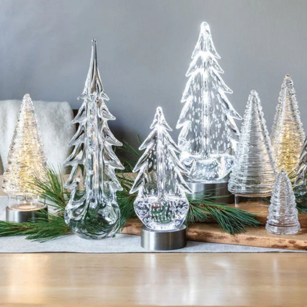 Clear glass evergreen trees on small and large LED illumination bases with a flat base and a central LED light, in a silver finish on a table with other holiday decor
