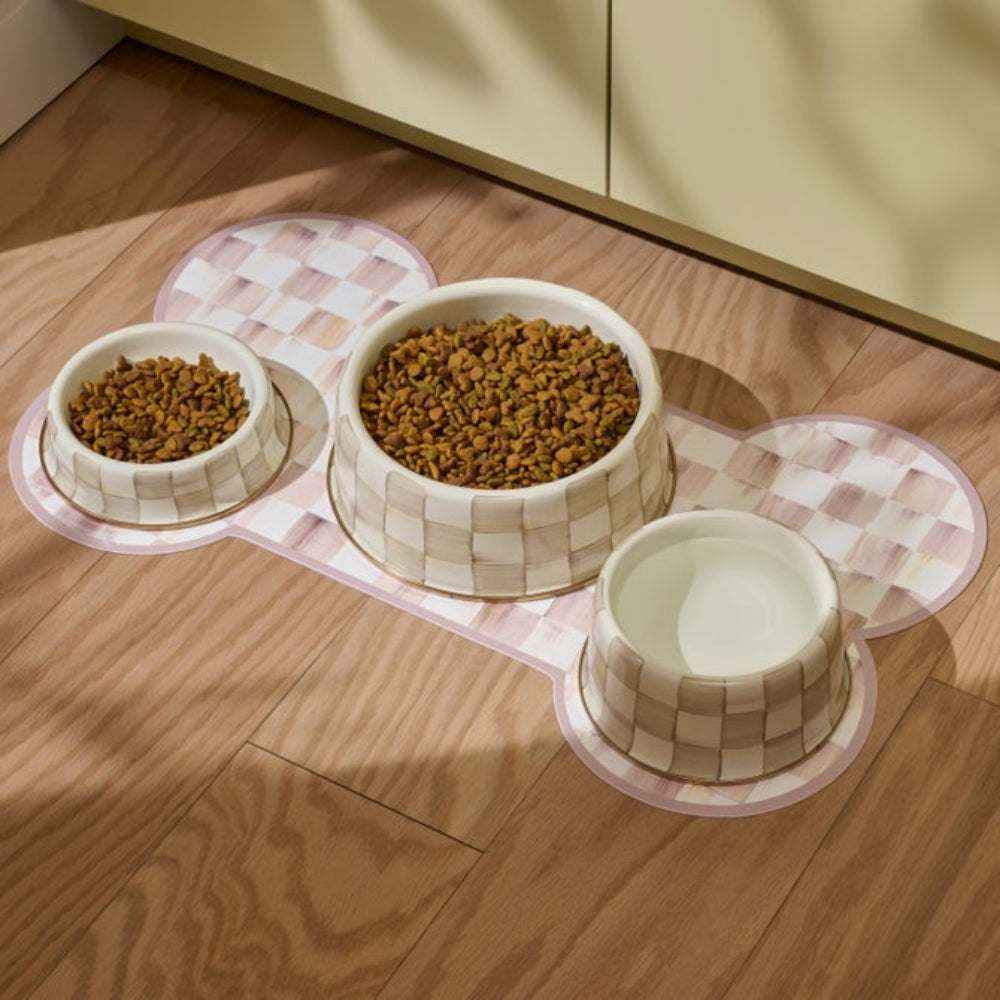 Three ceramic pet bowls with a beige and white checkered pattern sit on a matching bone-shaped mat on a wooden floor. Two bowls are filled with dry kibble, and one bowl is empty.