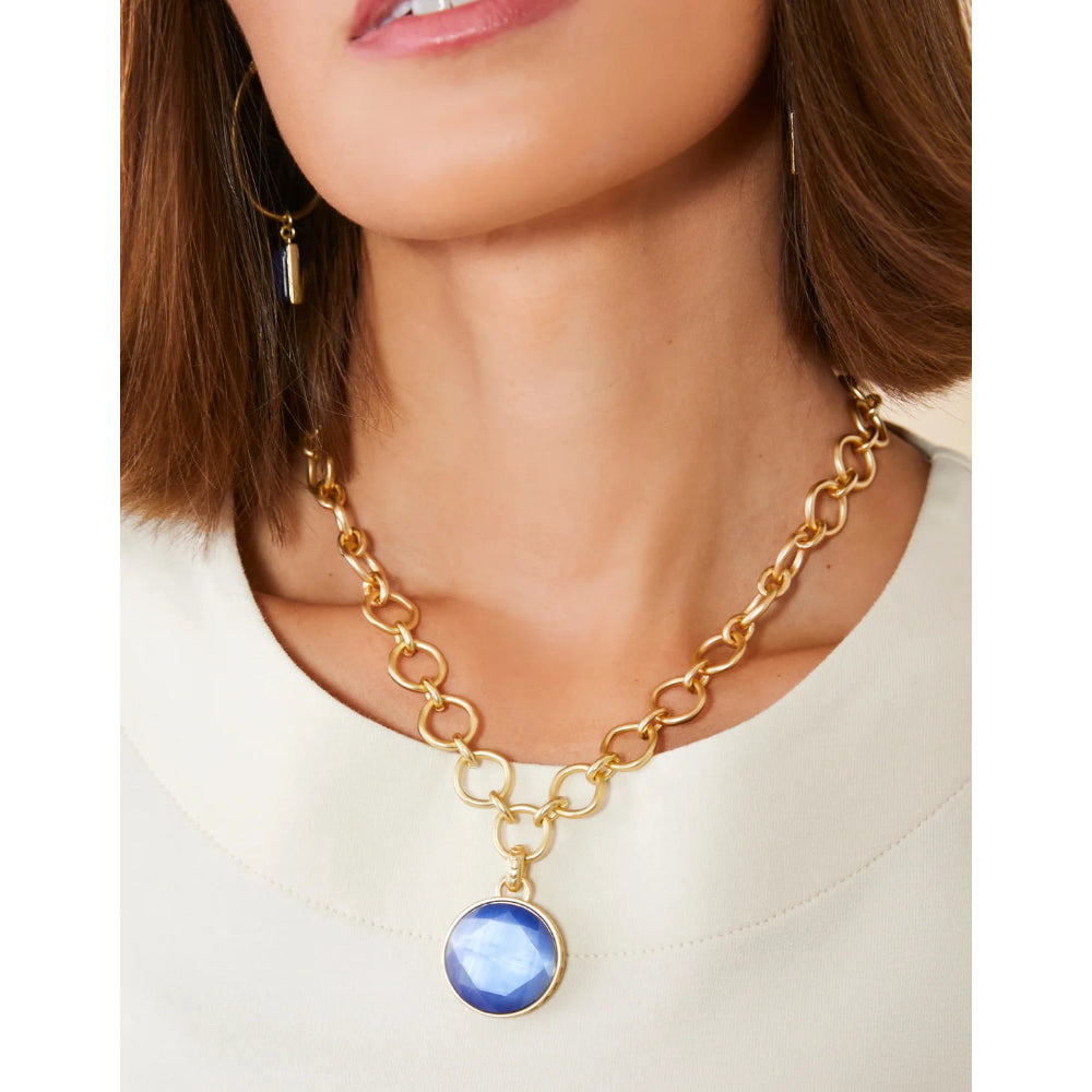 A close-up of a model wearing a gold chain necklace with a round blue gemstone pendant, styled with a cream-colored top.