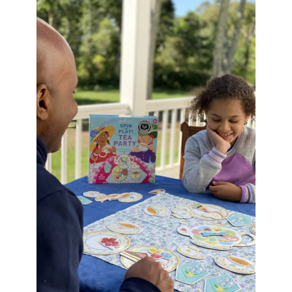 Two people playing a board game with a book titled 'Spin a Tea Party' on a table outdoors.