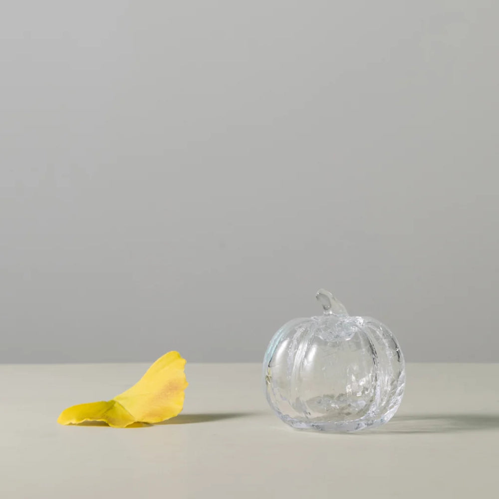 A clear glass pumpkin-shaped object with a small stem, placed on a light surface against a plain light gray background, with a yellow petal lying to the left.
