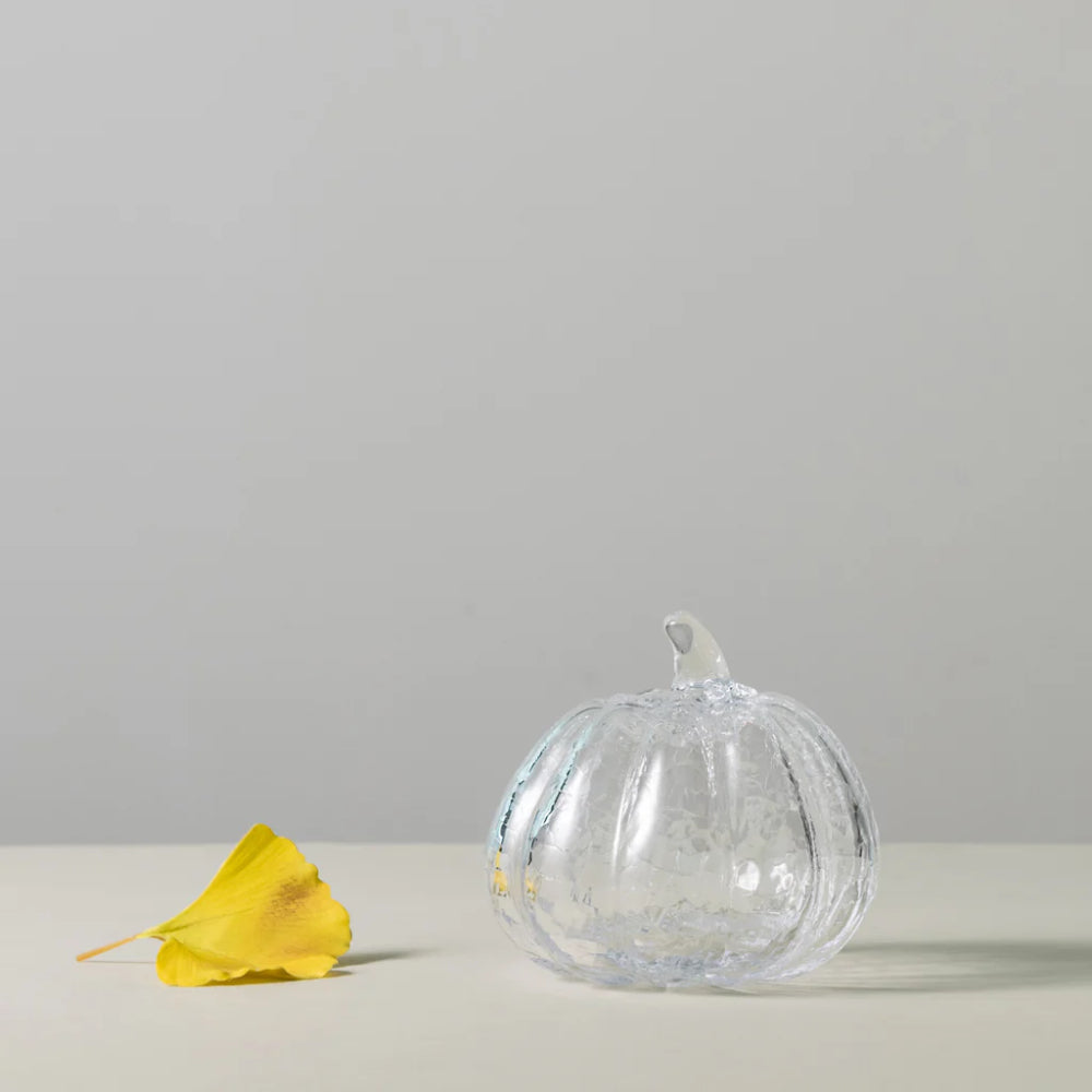 A clear glass pumpkin-shaped decorative object with a textured crackle finish sits on a white surface against a light gray background, with a yellow leaf placed to the left.